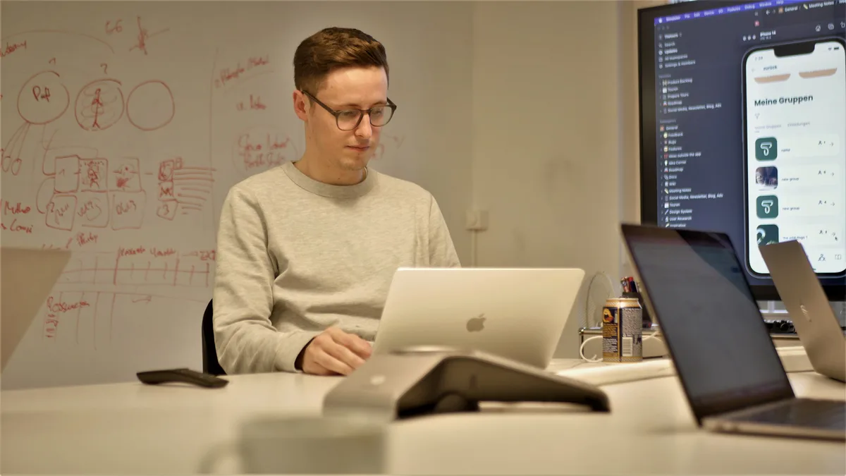 Man sieht einen jungen Mann in einem gläsernen Sitzungszimmer, der während einer Besprechung in seinen Laptop schaut. Neben den Laptop steht ein Mategetränk.